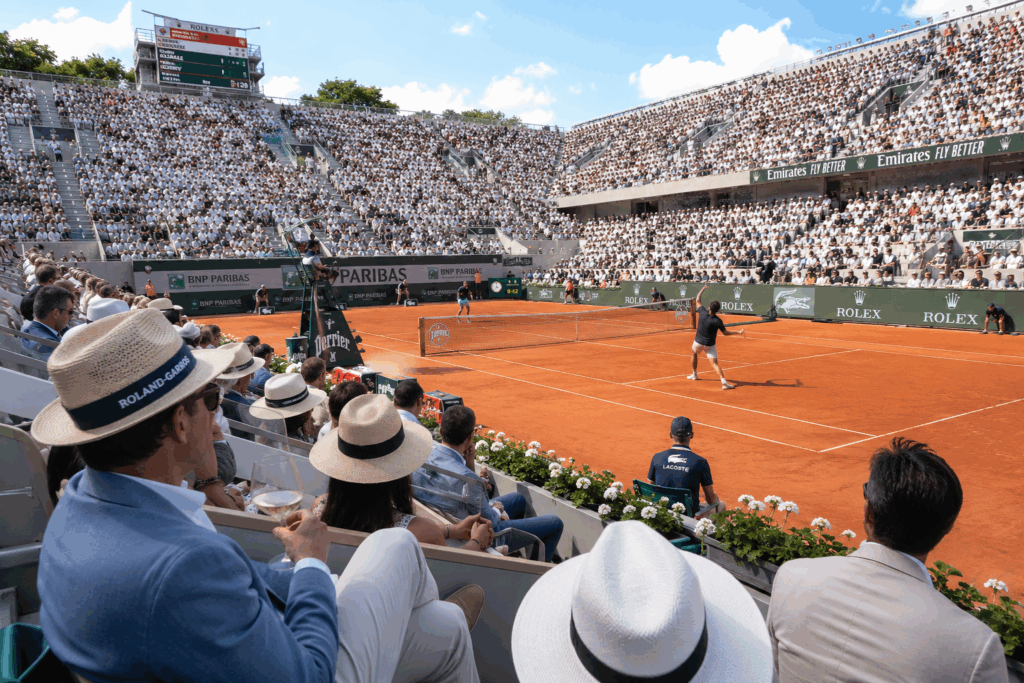 Spectateurs élégants assistant à un match de Roland-Garros 2026 depuis les gradins avec joueurs professionnels sur terre battue, arbitre et sponsors de luxe.