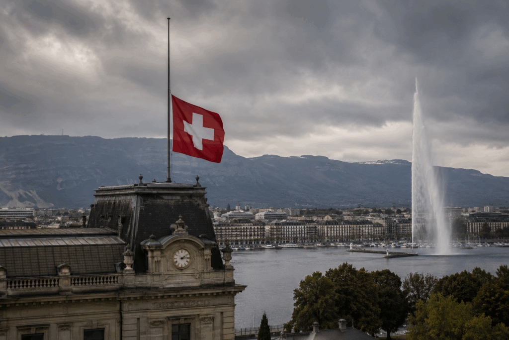 Drapeau suisse en berne au-dessus d’un bâtiment emblématique de Genève avec le Jet d’Eau en arrière-plan sous un ciel sombre