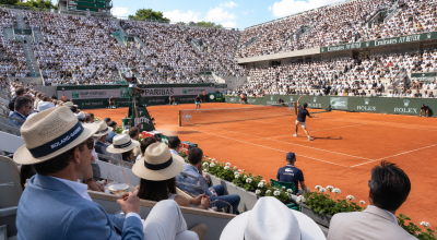 Spectateurs élégants assistant à un match de Roland-Garros 2026 depuis les gradins avec joueurs professionnels sur terre battue, arbitre et sponsors de luxe.
