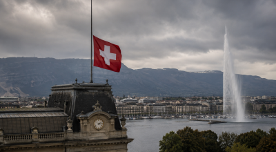 Drapeau suisse en berne au-dessus d’un bâtiment emblématique de Genève avec le Jet d’Eau en arrière-plan sous un ciel sombre